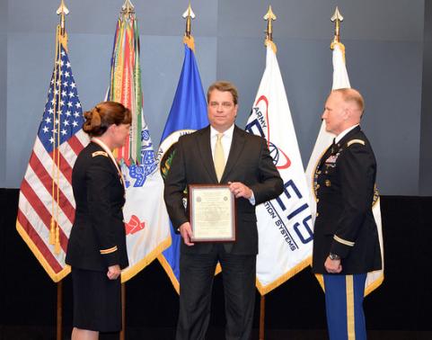 Two soldiers and a civilian stand on stage in front of flags. Civilian man is holding framed charter.