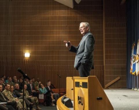 Dr. Bruce Jette speaks to a crowd while standing on a stage in an auditorium.