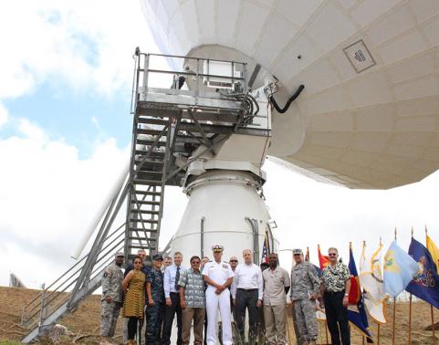 Group of civilians and soldiers stand around large satellite. 